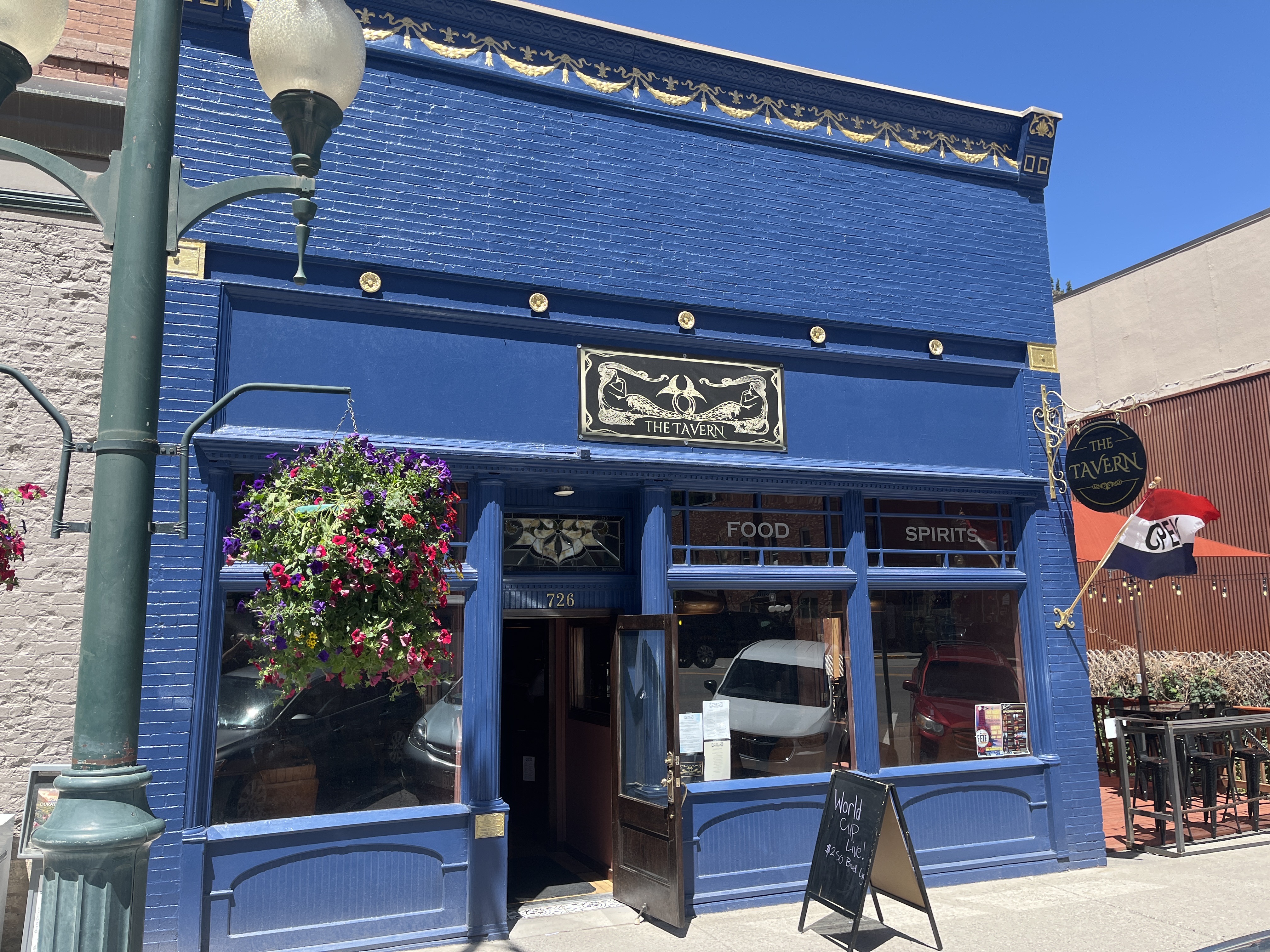 The Tavern storefront at 726 Main Street, Ouray — cobalt-blue brick facade with gilded relief, hanging flower baskets and FOOD · SPIRITS in the windows.