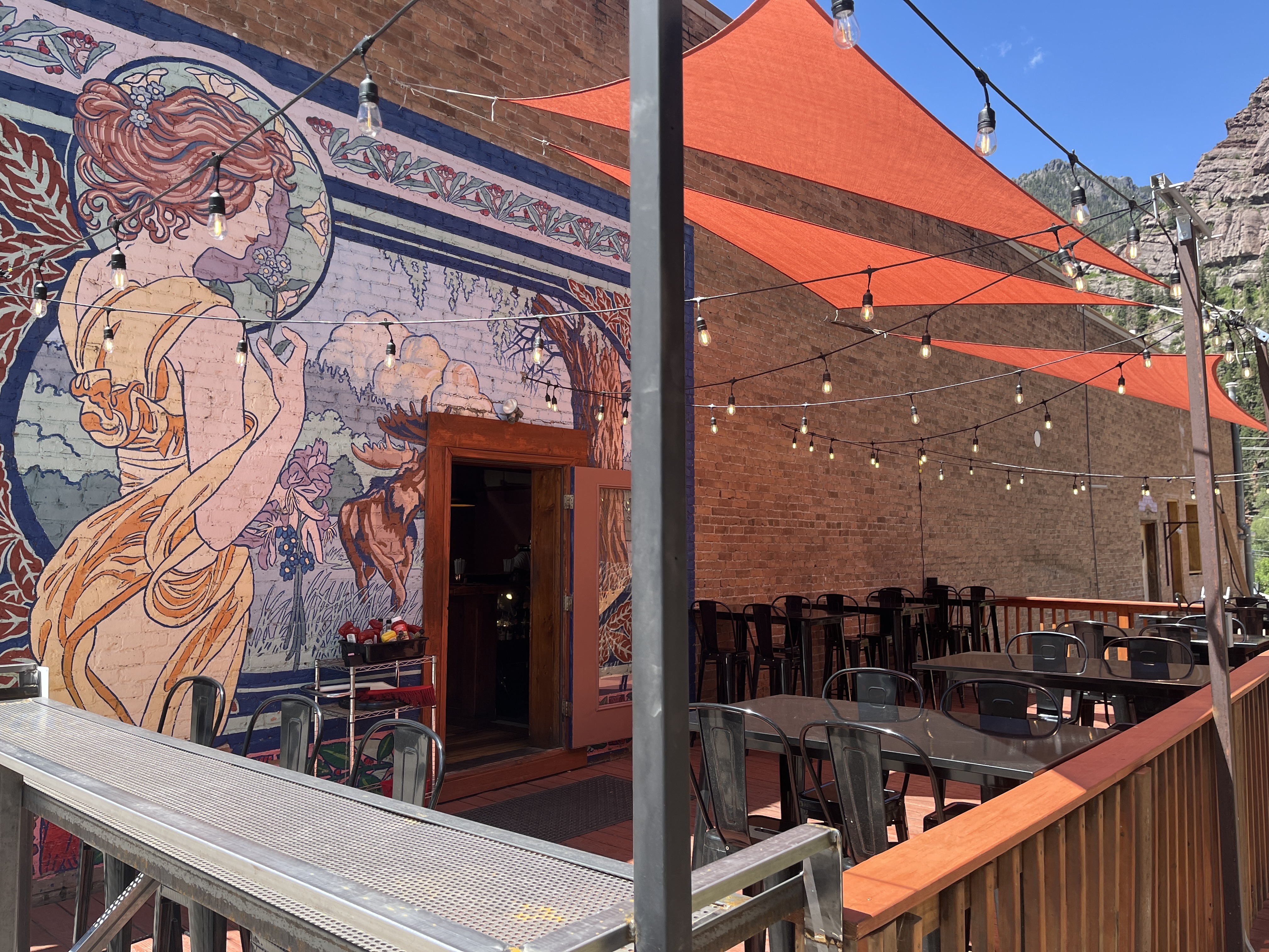 The Tavern outdoor patio with Art Nouveau mural, string lights and orange shade sails beneath the cliffs.